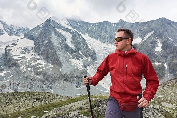 背景是一名男子穿着红色夹克，背着背包，在瑞士阿尔卑斯山脉的岩石岭上用登山杖散步