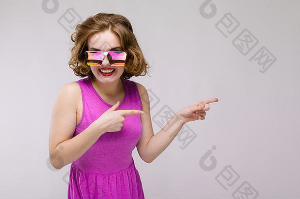 在灰色背景下穿着粉红色连衣裙的迷人年轻女孩。戴方眼镜的快乐女孩。那女孩用手指指向一边