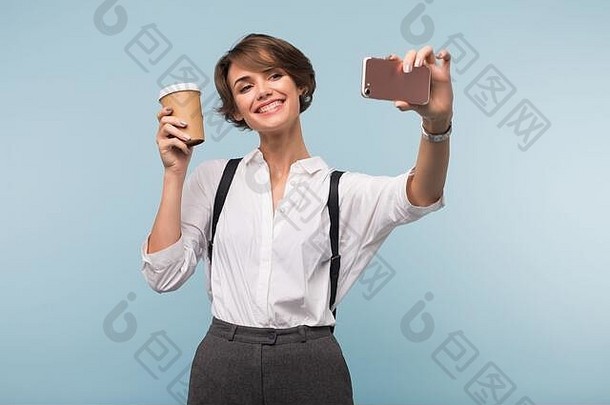 一位年轻开朗的女士，深色短发，身穿白衬衫，<strong>手持</strong>一杯咖啡在蓝色背景上愉快地用手机<strong>拍照</strong>