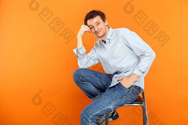 年轻的成人男人。坐椅子触碰头相机露出牙齿的微笑工作室拍摄橙色背景