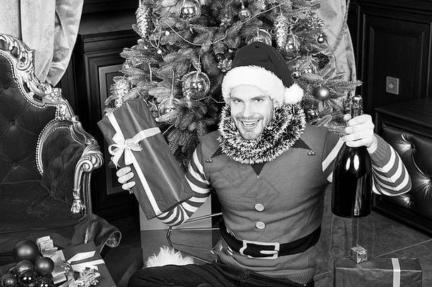圣诞树前，身穿精灵服装的圣诞老人微笑着拿着香槟瓶和礼品盒。庆祝，派对理念。新年快乐，圣诞快乐，节日快乐。送礼物，节礼日。