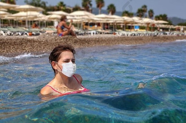 在阳光明媚的夏日，一位年轻的高加索妇女戴着医用防护面罩在海边游泳，背景是海岸线上的沙滩伞。假日