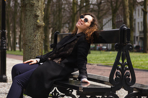 一个穿着黑色外套，留着长长的卷发，享受阳光的女孩。一位戴着黑眼镜的迷人年轻女子坐在春天公园的长椅上。