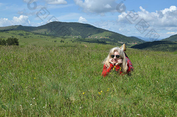 身着<strong>红色</strong>衣服的女人躺在山上的草地上休息，背景是美丽的风景