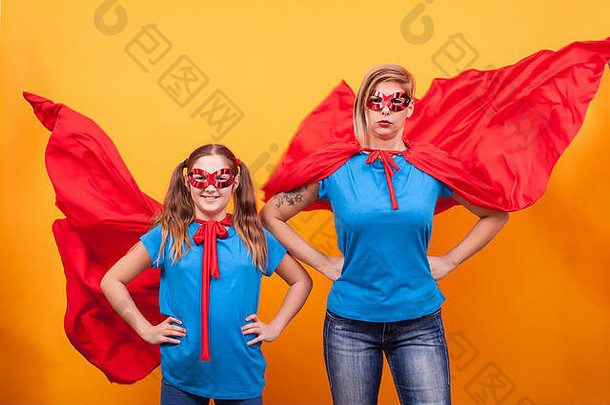 母女俩穿着超级英雄的服装，戴着红色的斗篷，在黄色的背景上飞翔。母女<strong>历险</strong>记。快乐的童年。