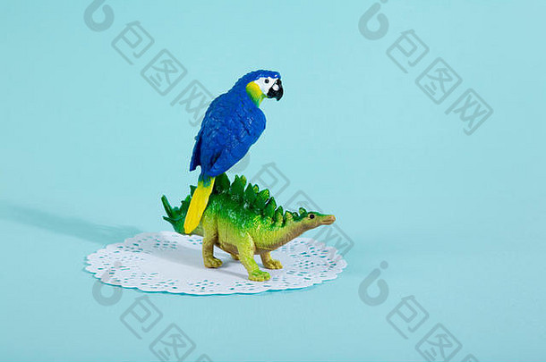 一只恐龙剑龙和一只蓝色鹦鹉站在一张白色蕾丝纸上，背景是充满活力的绿松石色。极小的非拍静物摄影