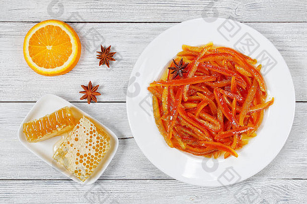 木桌上的白色盘子上放着美味的蜜饯橘子和柠檬皮，背景是蜂蜜和半个橘子，从上面看
