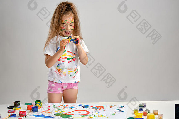 穿着白色t恤的小女孩站在桌旁，手里拿着惠特曼和五颜六色的颜料，玩着浸在颜料里的海绵。白色的隔离带。特写。