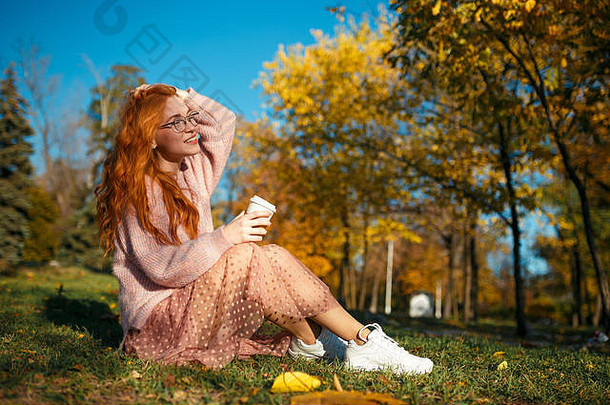 一个迷人的红发女孩的肖像，戴着眼镜，一张漂亮的脸。一个穿着毛衣和珊瑚色裙子的女孩在秋天公园摆姿势。在少女时代