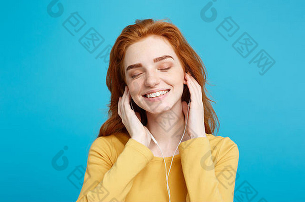生活方式理念——欢快快乐的姜红色头发女孩的肖像，戴着耳机欣赏音乐，对着镜头欢快地微笑。在蓝色粉彩背景上隔离。空间。