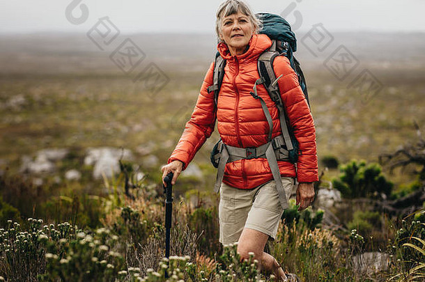 徒步旅行中富有冒险精神的老妇人。穿着夹克和背包，手持登山杖在乡村徒步旅行的老妇人。