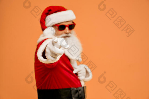 酷炫时尚的成熟圣诞老人，戴着墨镜和白手套，留着真正的灰色胡须，手指指向相机，背景是模糊的橙色。正面积极自信的姿态。