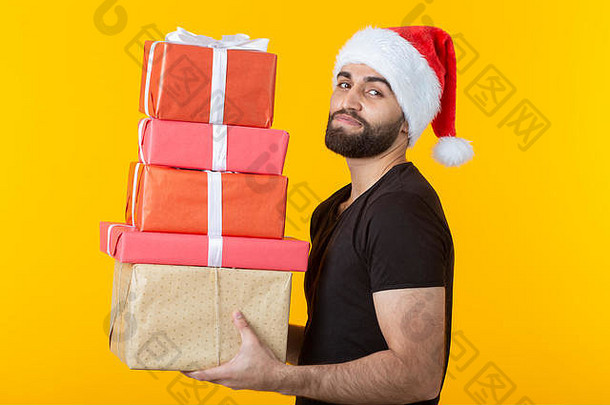 戴着圣诞老人帽子、留着胡子、心怀不满的年轻人拿着五个礼盒，在黄色背景上摆姿势。礼物和问候的概念