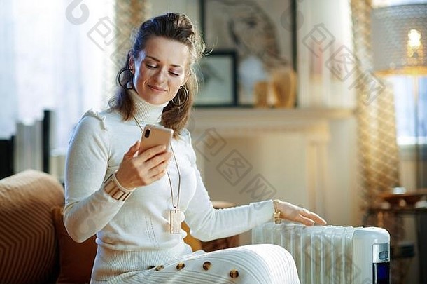 在阳光明媚的冬日，一位穿着白色毛衣和裙子的优雅女士坐在沙发上，坐在白色电油散热器旁，用智能手机应用程序为自己设计手机