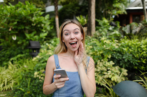 一位穿着牛仔裤和太阳裙的年轻快活女人在树旁的花园里用智能手机聊天。