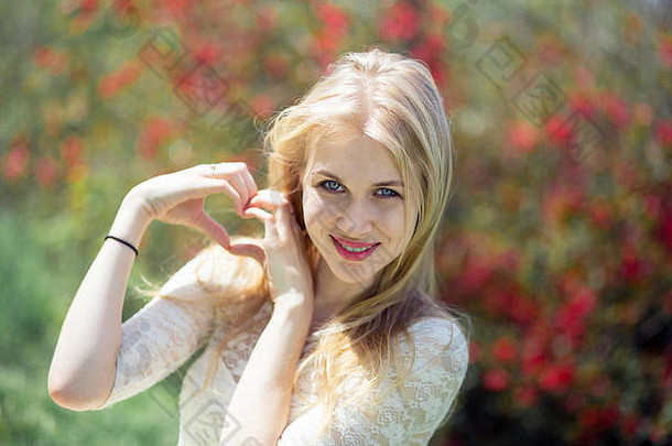 姿态爱自然概念-微笑的金发女郎在花开的春天背景上展示手指做成的心形