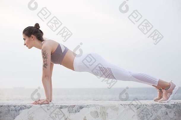 穿着<strong>运动</strong>上衣和白色紧身裤的美丽女孩练习瑜伽，背景是海景。年轻女子独自在海边梦幻般地训练