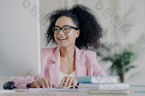 快乐的卷发职业女经理的照片聚焦在电脑屏幕上，笑容灿烂，卷发，戴着透明眼镜，穿着优雅的西装，