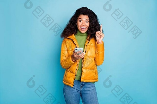 时髦的深色皮肤卷曲的女士拿着电话举起食指有酷的创意帖子思想穿黄色外套牛仔裤绿色毛衣的照片