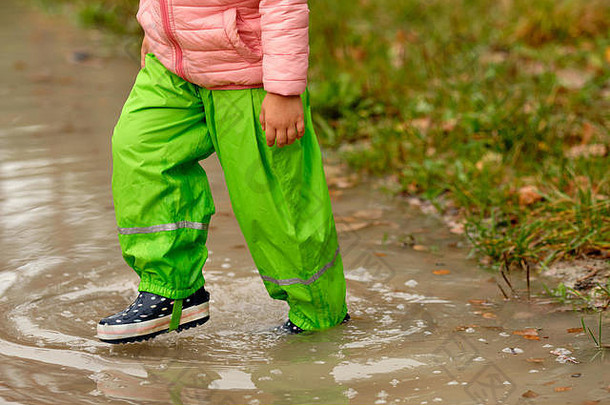 十月一个秋天的雨天，一个穿着绿色防水裤和橡胶靴的小女孩在森林里走过一个巨大的雨水坑