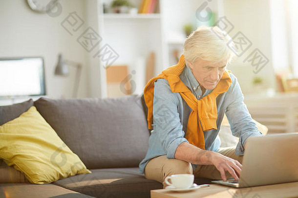 现代老人在家工作时使用笔记本电脑的肖像，坐在客厅的沙发上，复印空间