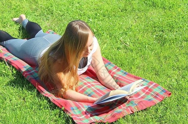 一个穿着白色T恤、留着长发的年轻漂亮的白人女孩躺在一块红色格子布上，躺在绿草上，躺在草坪上看书。
