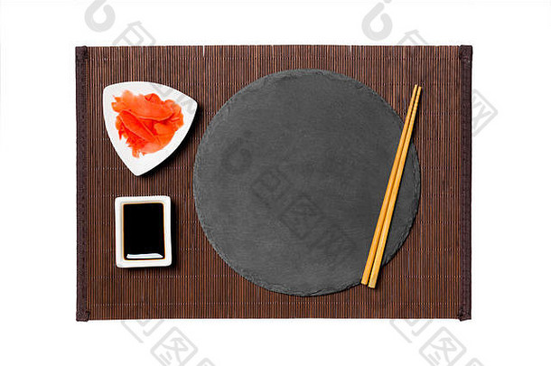 在深色竹垫背景上，用筷子清空黑色板岩盘子，盛寿司、生姜和酱油。带有供您设计的空间的俯视图。