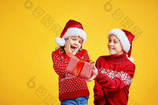 戴圣诞帽的快乐男孩将圣诞礼品盒送给穿着红色毛衣、背景为黄色的鹿的兴奋女孩