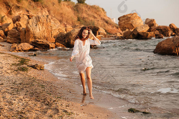 身着浅色夏装、面带微笑的黑发女子在海边散步的全幅照片