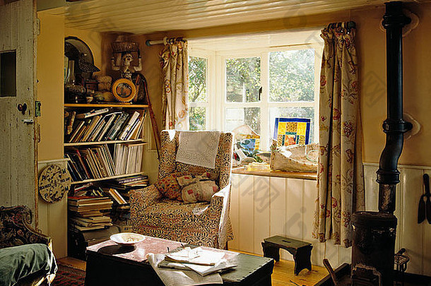 低矮平房客厅窗户前的扶手椅，有凌乱的书架和烧木头的小火炉