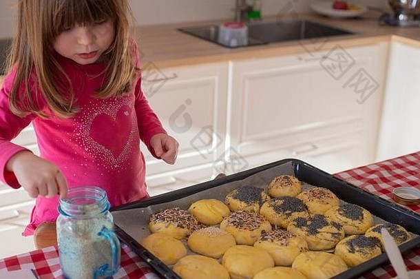 高加索女孩，3岁，在烹调前在烤箱里的面包包子上撒种子。封锁活动的想法。
