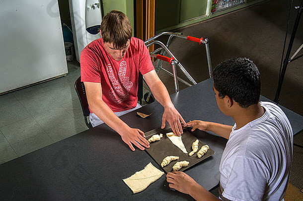 在烹饪课上，一位老师帮助一位视力受损的残疾青少年用准备好的面团通过触摸来制作面包卷。