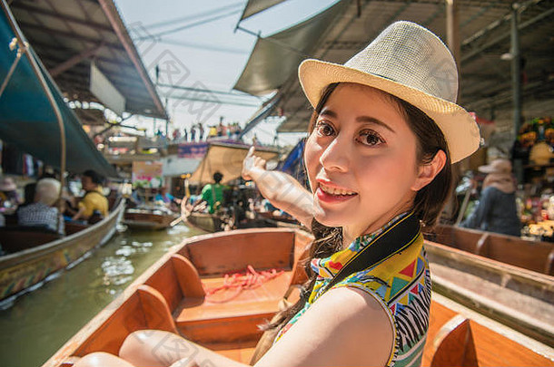 在泰国旅游期间，微笑的美女旅游妇女指着当地小贩的风景为朋友分享，坐在河船上参观著名的达姆诺