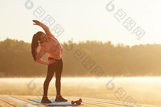 一个正在训练的小女孩在秋天日出时在湖边蹲着。