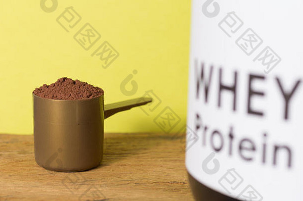 用于训练和运动的乳清蛋白食品补充剂。木桌上放着巧克力粉和黑罐子的勺子的特写镜头。颜色背景：耶洛