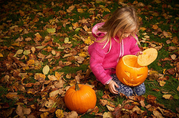 可爱的金发女孩在秋天的树叶上玩万圣节<strong>南瓜</strong>。