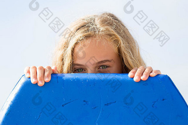 一个厚颜无耻的年轻女孩躲在一块冲浪板后面，在海滩上享受夏日的阳光。