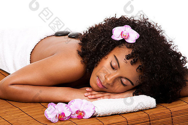 <strong>健康</strong>日水疗中心的快乐女人用白色毛巾铺在装饰着兰花的竹桌上，接受热石按摩治疗。