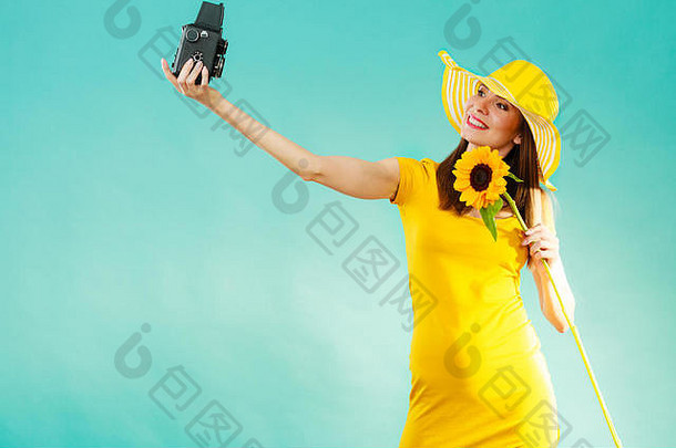 夏天的女人穿着黄色的裙子，戴着向日葵的帽子，在生动的蓝色背景下用老式相机拍摄自己的照片
