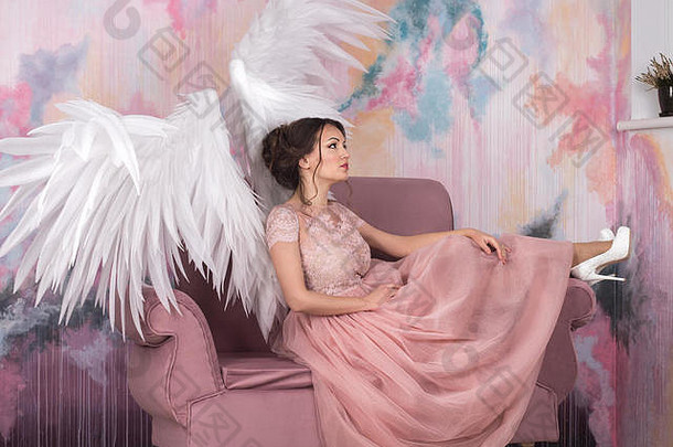 美丽的年轻模特，张开天使的翅膀坐在工作室的沙发上。白色背景