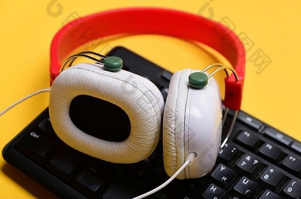 带有电脑键盘的红白两色耳机。录音和技术。耳机和黑色键盘。暖黄色背景上的电子设备。音乐和呼叫中心概念