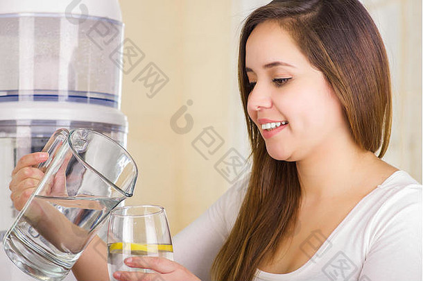 美丽的女人一手拿着一杯水，另一手拿着一罐水，厨房背景上有一个净水器过滤系统