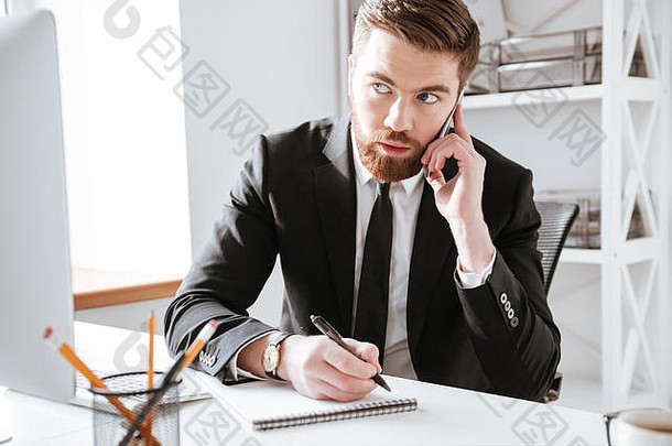 一张专注的商人坐在办公室边打电话边写笔记的照片。