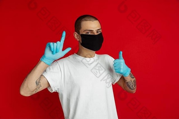 在红色工作室背景下，一名戴着防护面罩和医用手套的白人年轻人竖起大拇指，指指点点。人类情感的概念，销售，冠状病毒锁定，广告，隔离，绝缘。