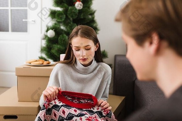一幅美丽女孩的画像，她拿着新毛衣坐在家里的地板上，若有所思地看着它，背景是圣诞树