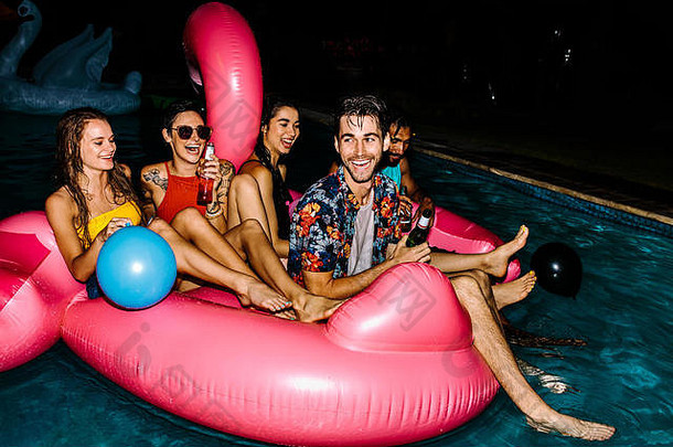 一群男女在一个充气的火烈鸟游泳池浮动床垫上欢笑。快乐的朋友们晚上在游泳池里闲逛。埃维尼
