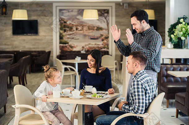 家庭、亲子关系、科技、人的概念——快乐的母亲、父亲和小女孩正在吃饭，请服务员用智能手机拍照