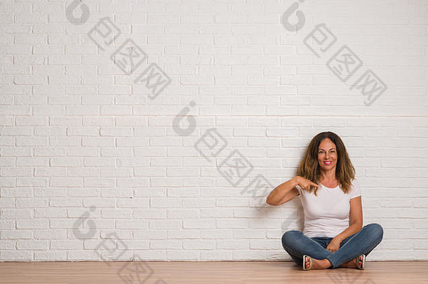 中年西班牙裔妇女站在白砖墙上，惊讶地用手指着自己