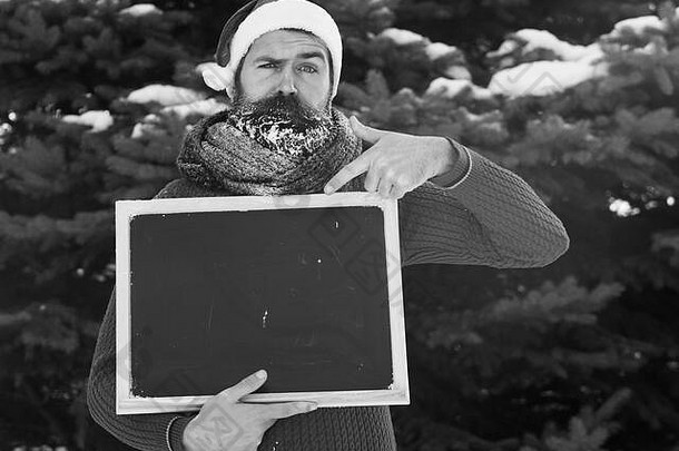 戴着圣诞老人帽子的皱眉头的男人，留着胡子、留着白霜胡子的时髦男人，在冬天的自然背景上指着空白的黑板或黑板，空间
