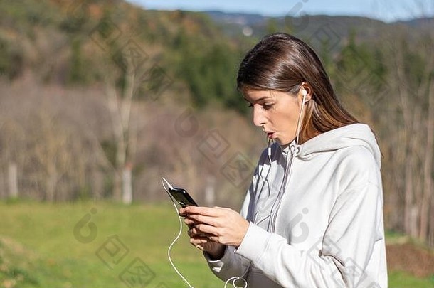 在一个阳光明媚的日子里，一位漂亮、惊讶的黑发女人在公园里用智能手机阅读新闻。她穿着白色毛衣，留着长发。